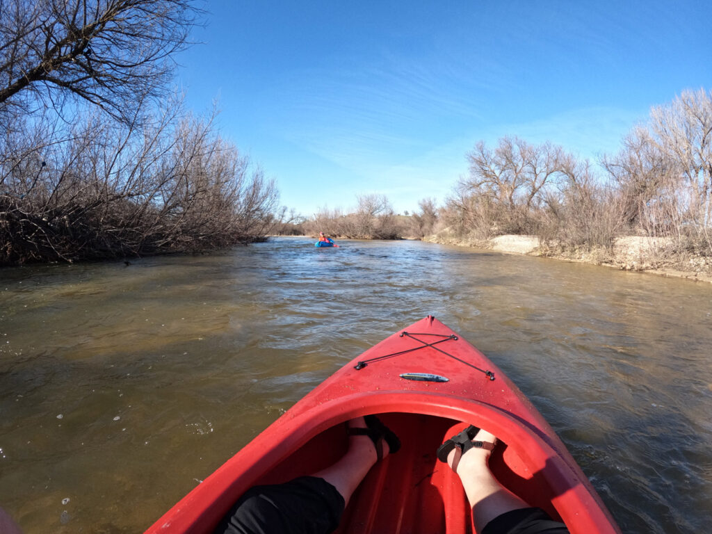 View from the front of a kayak floating down a deeper section of river. Another person in a packraft is visible up ahead.