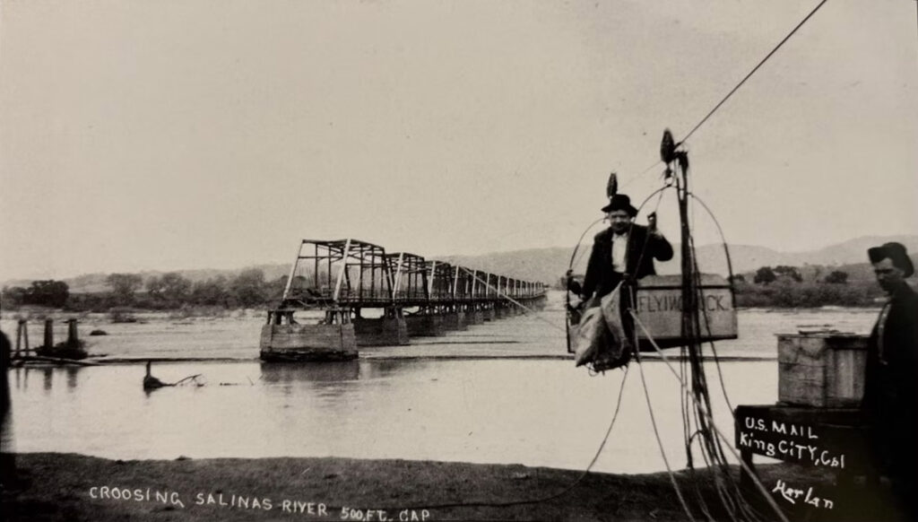 Historic photo of the King City Bridge over the Salinas river in the early 1900s. A man sits in a "flying duck" tool that transports him to the bridge over the water.